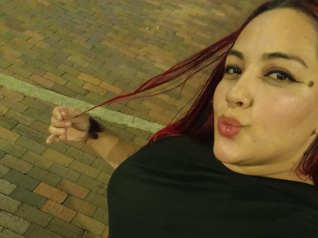 LaGorditaPelirRoja mit roten Haaren, schwarzem Hemd, leicht geschürzten Lippen, die Haare haltend, braunen Augen, einem kleinen Schönheitsfleck auf der Wange, stehend auf einem Ziegelboden, leicht übergewichtig, süß..
