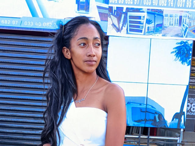 Diamond, une femme aux longs cheveux noirs, à la peau brune et avec de petites seins, portant une robe blanche sans bretelles, se tient devant un mur bleu et blanc réfléchissant, et regarde sur le côté..