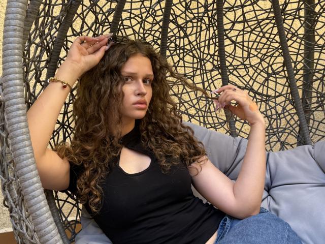 CurlyElina, con cabello castaño rizado, un top corto negro que muestra sus pechos, jeans azules, sentada en una silla de mimbre gris, con la mano en el cabello, con una expresión seria, y llevando una pulsera dorada..