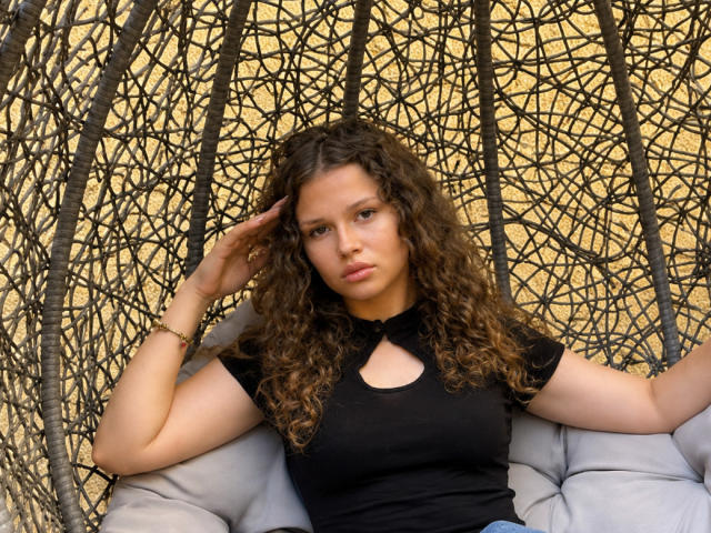 CurlyElina, con i capelli castano ricci, una maglietta nera e piccoli seni, seduta su una sedia di vimini, con una mano tra i capelli, che appare seria, indossa un braccialetto, su un cuscino beige..