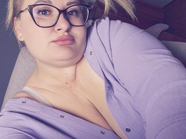 RadaRori with glasses, light purple shirt, showing cleavage, white bra strap visible, pouty lips, blonde hair, slightly chubby, fair skin, seated, taking selfie, subtle nose ring.