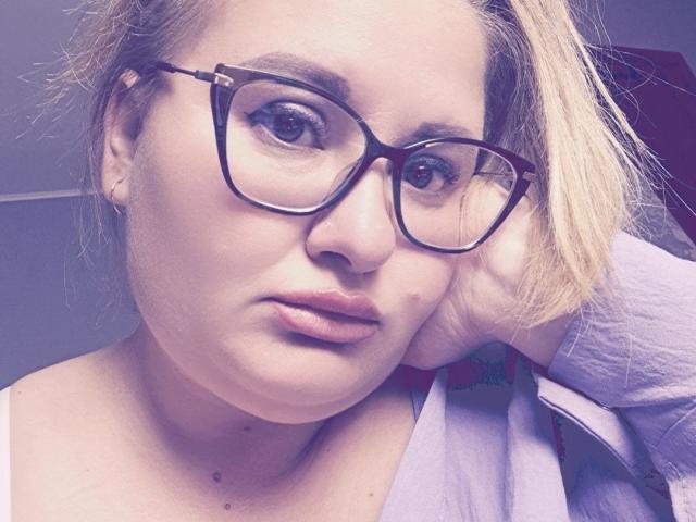 RadaRori mit Brille, voller Lippen, blonden Haaren, heller Haut und kleinen Sommersprossen, trägt ein weißes Hemd, sieht ernst aus und hält sich ihre Wange..