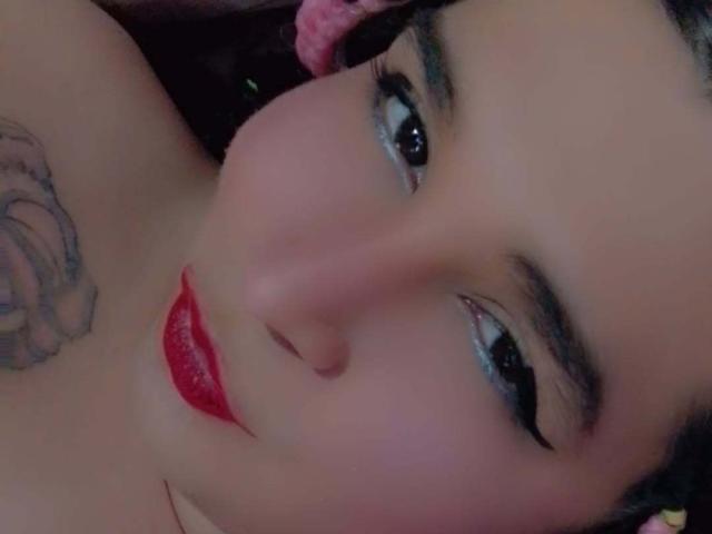 Keissydoll hat ein süßes Gesicht mit rotem Lippenstift, schwarzem Eyeliner und einem Rosentattoo auf ihrer Schulter. Ihr dunkles Haar und ihre glänzenden Augen sind auffällig..