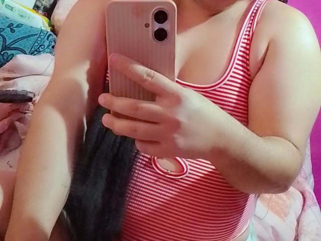 Keissydoll with pink-striped tank top, holding phone, showing her tits and arm, black hair in background, pink phone case, pink wall, pink bed, slightly visible ass.