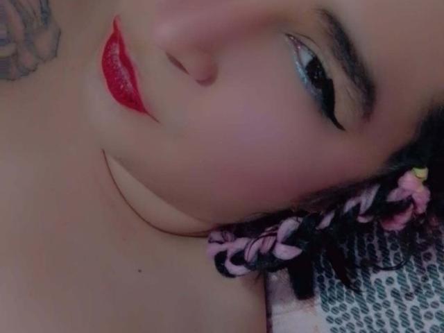 Il viso di Keissydoll, labbra rosse e lucide, eyeliner nero, capelli rosa intrecciati con fiori, tatuaggio sul lato sinistro, pelle rosa pallida, sdraiata, leggermente sfocata..