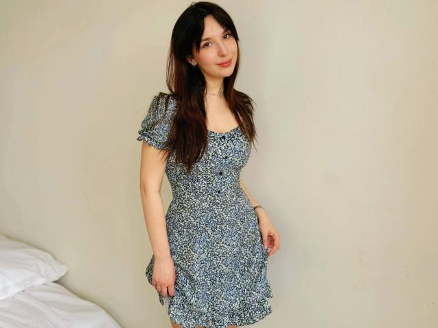 DirtyAngel: una chica atractiva con cabello castaño largo, un vestido azul estampado, pechos pequeños y una sonrisa encantadora, de pie junto a una cama, mirando ligeramente hacia un lado..