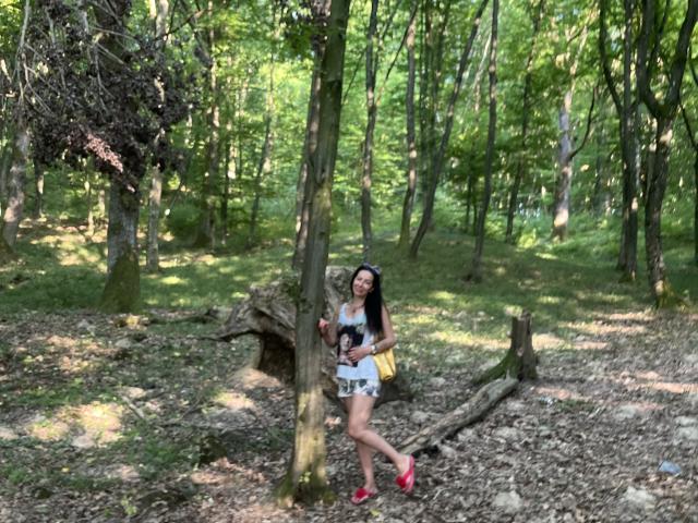 MirandaHott69 con cabello largo y negro, camiseta blanca, pantalones cortos amarillos, zapatos rojos, de pie junto a un árbol en un bosque, sosteniendo una cámara, con un bolso amarillo pequeño..