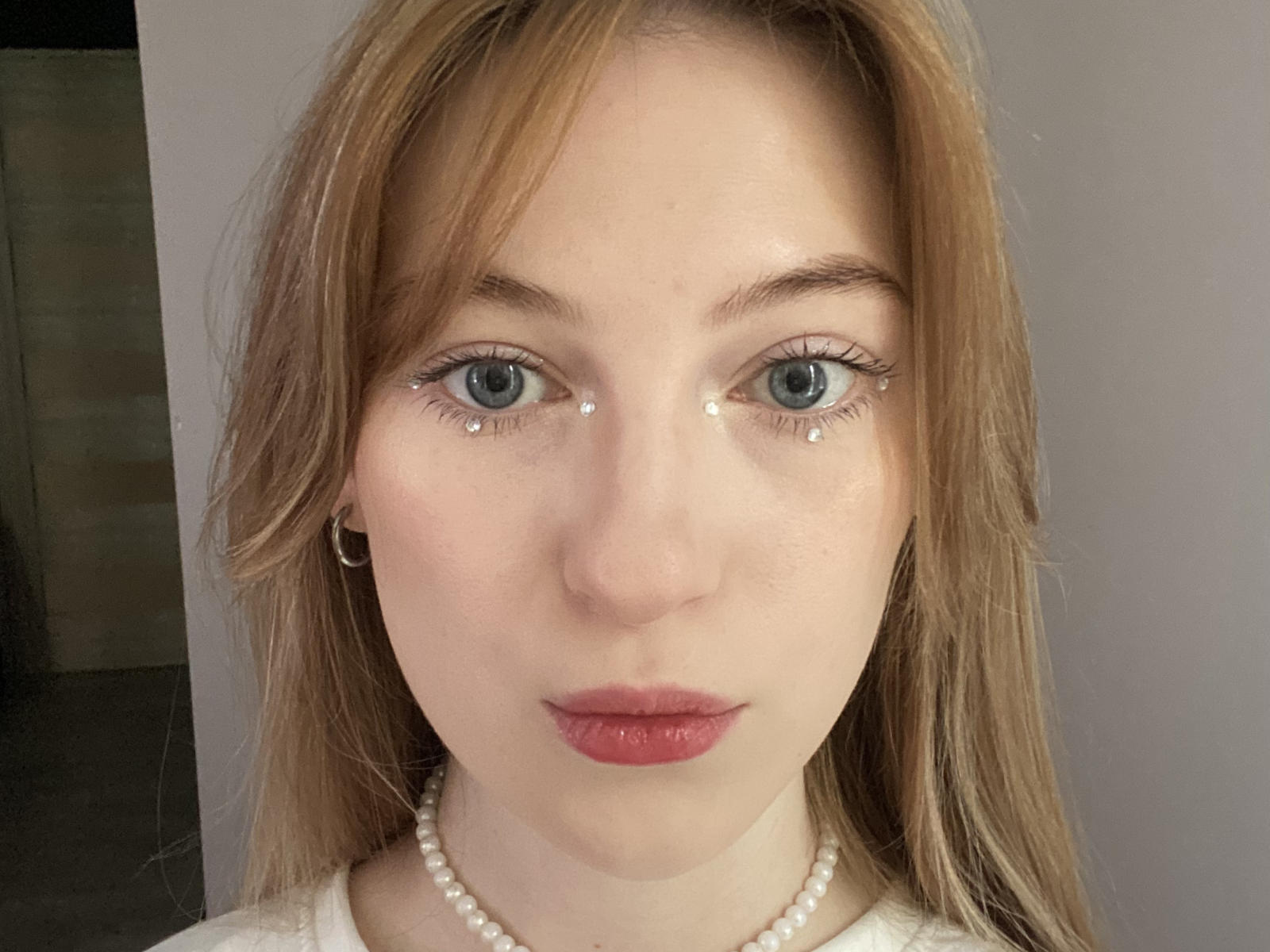 ZettaGipe tem olhos azuis com pequenas joias em forma de diamante, pele clara, batom vermelho, cabelos longos loiros e um colar de pérolas brancas, usando um pequeno brinco de argola dourado..