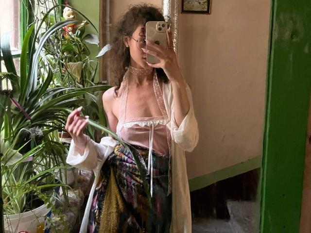 AlyceErway with curly brown hair, glasses, pink choker, sheer white top, floral skirt, holding green plant, mirror selfie, phone in hand, reflection, green door frame, plants in background.
