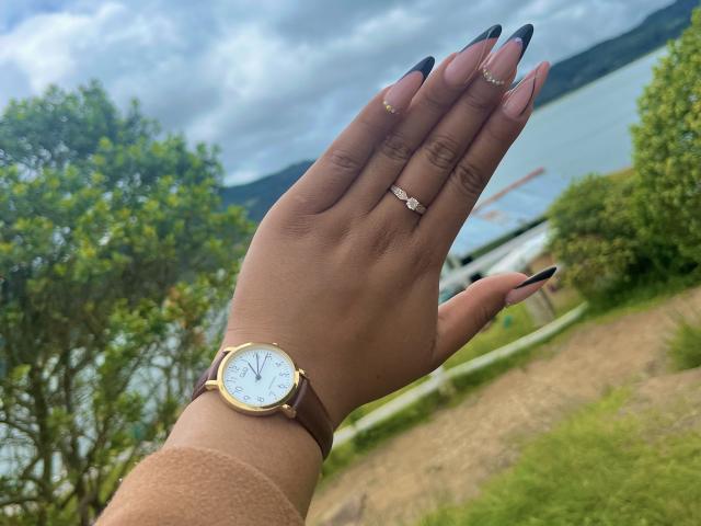 A mão bronzeada de SophieSonyas, com unhas longas e pretas, um relógio de ouro, um anel e um anel de diamantes, contrastando com um céu nublado e uma paisagem verde..