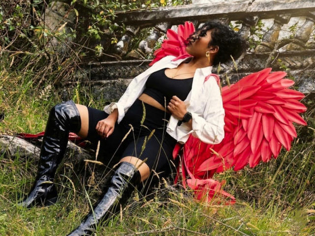 SophieSonyas con grandes senos, vestido negro, chaqueta blanca, botas negras brillantes y alas de ángel rojas, sentadas en el césped, mirando hacia arriba con cabello negro rizado, al aire libre..