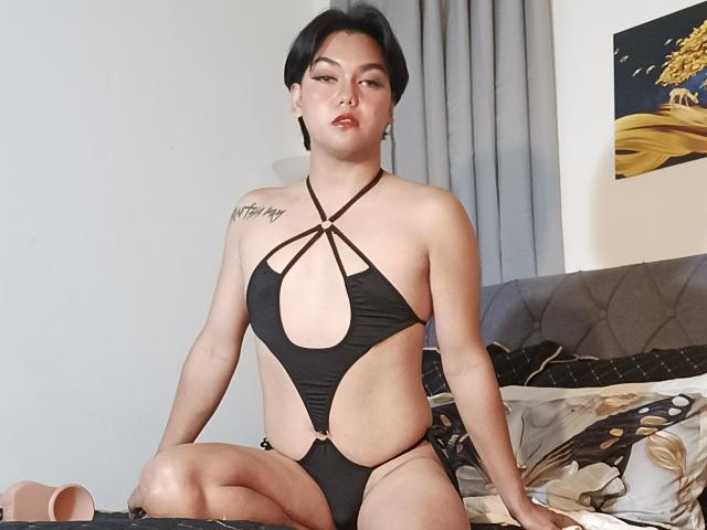 Ein FemboyMnl mit kurzen, schwarzen Haaren, der ein freizügiges, schwarzes Einteiler-Top trägt, das Brüste und Vulva zeigt, sitzt auf einem Bett, blickt in die Kamera und hat ein Tattoo an der rechten Schulter..