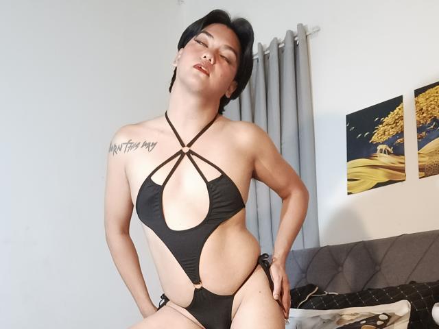 FemboyMnl con capelli neri corti, che indossa un monokini nero con spalline sottili e scollato, mettendo in mostra un seno abbondante e un tatuaggio sul lato destro del petto, che posa con sicurezza, con gli occhi chiusi e le labbra leggermente aperte..