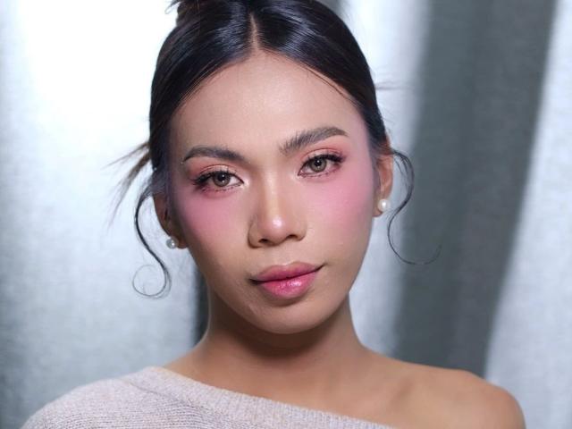Brittany08-hot tiene un rubor rosado en las mejillas, ojos marrones y labios rosados brillantes. Lleva una blusa beige con un hombro descubierto y aretes de perlas, con su cabello negro recogido en un moño..