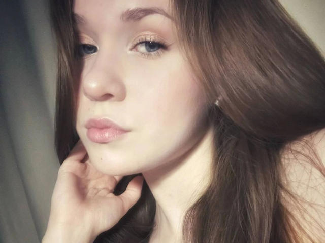 Kristty-hot tiene piel clara, cabello largo y castaño, ojos azules y labios rosados. Está mirando hacia abajo con la mano cerca de la barbilla, mostrando una pequeña peca en la mejilla..