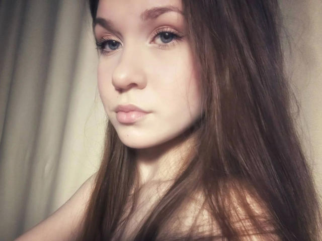 Kristty-hot hat eine blasse Haut, blaue Augen und langes, braunes Haar. Ihre rosafarbenen Lippen sind leicht geschürzt. Sie blickt mit einem neutralen Ausdruck in die Kamera. Ihre Brüste sind nicht sichtbar..