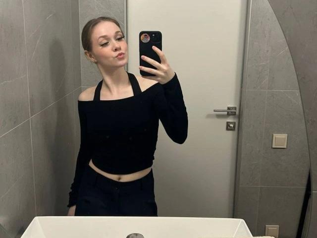 Kristty macht ein Selfie vor dem Spiegel, trägt ein schwarzes Top mit einem Schulterauschnitt und schwarze Hosen, hält ein Handy in ihrer rechten Hand, zieht die Lippen zusammen und befindet sich in einem Badezimmer..