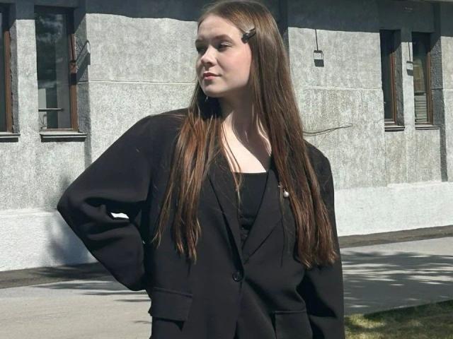Kristty, mit langen braunen Haaren, einer schwarzen Jacke und einem schwarzen Oberteil, steht mit den Händen in den Hüften, blickt zur Seite, hat eine kleine Haarnadel im Haar und ein ernstes Gesicht..
