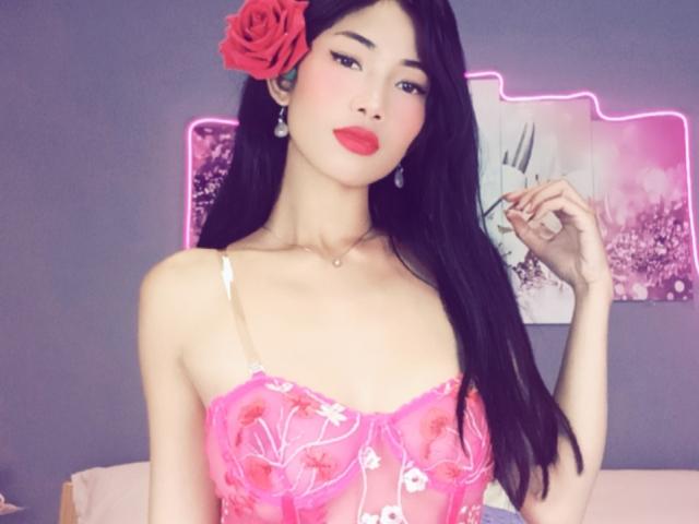 SweetPinkieFrench con cabello negro largo, una rosa roja en el cabello, lencería de encaje rosa con bordados florales, lápiz labial rojo y un collar, de pie, con la mano levantada, mirando a la cámara..