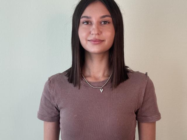 LangCarpinello, com cabelo castanho reto, pele marrom média, seios pequenos, vestindo uma camisa marrom e um colar de prata, sorrindo discretamente, em pé contra um fundo simples..