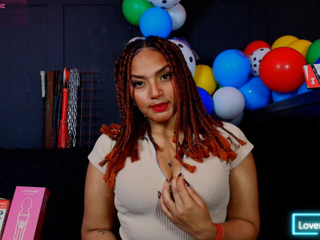 BrianaPratt met roodbruin gevlochten haar, die een beige geribbelde top draagt, haar borst aanraakt en glimlacht, omringd door kleurrijke ballonnen, heeft een gemiddelde borstmaat en draagt rode lippenstift..