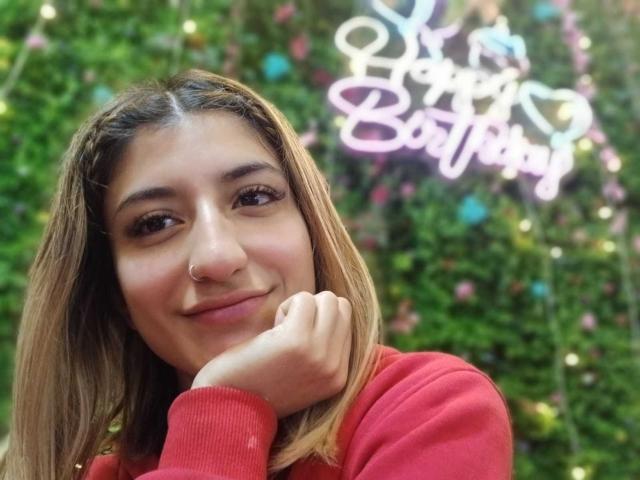 Kaiix, con cabello castaño, suéter rojo, piercing en la nariz, sonriendo, con la mano en la mejilla, mirando a la cámara, letrero de neón que dice "Feliz Cumpleaños" en el fondo borroso, pared verde con pequeñas decoraciones coloridas..