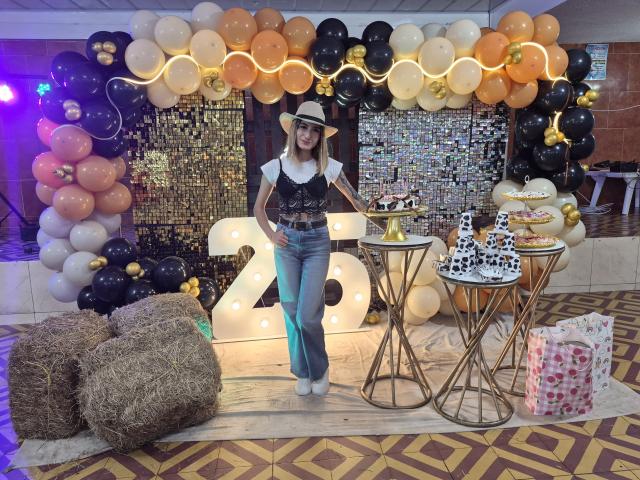 Kaiix está de pie con las manos en las caderas, usando un sombrero blanco, un chaleco negro y pantalones vaqueros azules, rodeado de globos, fardos de heno, malla dorada y un cartel con el número "2" en dorado..