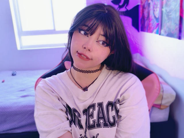 Arieyaara mit langem, schwarzem Haar, Pony und einer schwarzen Halskette, trägt ein weißes "METACASE"-Shirt und sitzt auf einem rosa Stuhl, wobei sie leicht lächelnd zur Seite schaut..