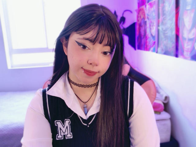 Arieyaara mit langen braunen Haaren, weißem Hemd, schwarzer Weste, einem "M"-Abzeichen, einem Choker, einem Nasenring und pinkem Lippenstift, steht in einem Raum mit violetter Beleuchtung. Brüste sind unter der Weste sichtbar..