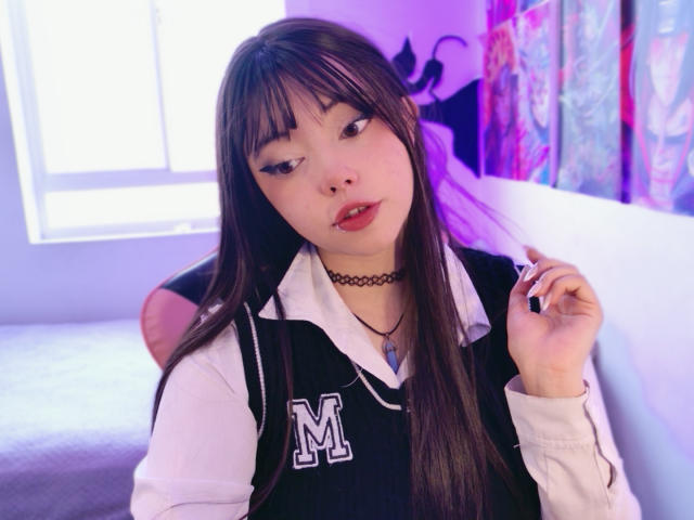 Arieyaara mit langen, braunen Haaren, Pony und einer schwarzen Halskette, trägt eine schwarz-weiße Sportjacke mit einem "M"-Logo, beißt sich auf den Nagel und schaut in die Kamera..