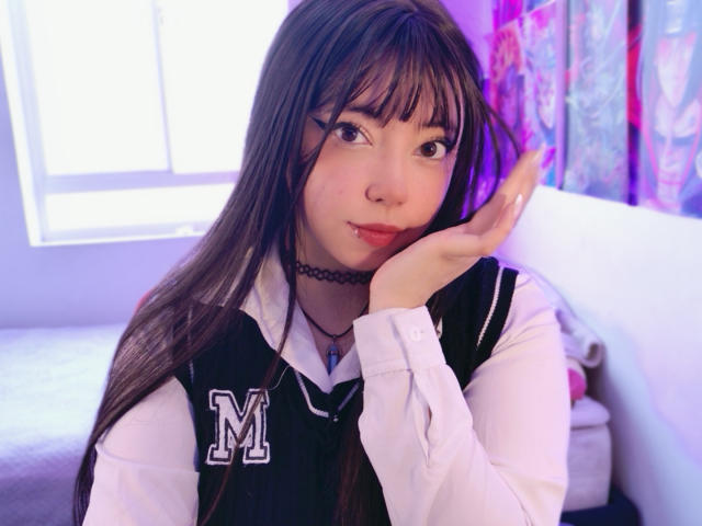 Arieyaara con lunghi capelli castani, frangia, collana nera, camicia bianca, gilet nero con la lettera "M", mano vicino al viso, luce viola, carina, piccolo piercing al naso, labbra rosa, che guarda la fotocamera..