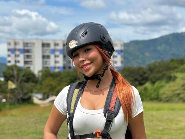 VaniaRizzo, com cabelo laranja, capacete preto, camisa branca e arnês, sorrindo, mostrando os seios, em pé em uma área gramada com montanhas e um prédio ao fundo..