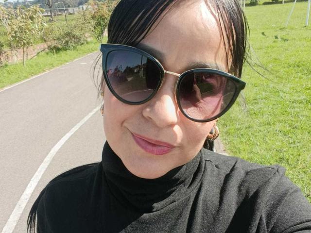 TamaraFoxy with black turtleneck, wearing dark sunglasses, pink lipstick, and black hair, smiling slightly, standing on a sunny grassy path, her ass and tits not visible.