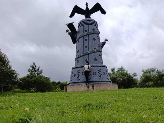 TamaraFoxy stands in front of a tall, dark statue with a bird on top, arms raised. She's wearing a white shirt and dark pants, with green grass around.
