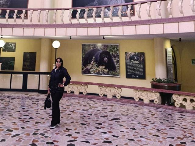 TamaraFoxy stands in a tiled, multi-level room, wearing glasses, black jacket, and pants, with a hand on her hip, holding a bag, under a white globe light.