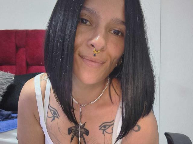 AmelieBlake with black shoulder-length hair, smiling, nose ring, white tank top, tattoos on chest, necklace, white walls, red headboard, black and gray pillows in background.