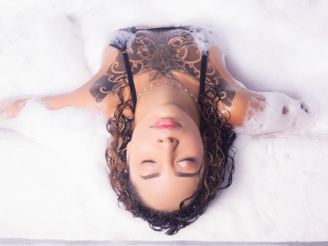SilvanaXOwen lying in a bathtub full of white bubbles, eyes closed, black tattoo on chest, curly brown hair, pink lipstick, necklace, water and bubbles around her shoulders.