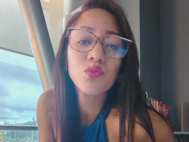 SamanthaCorine, avec ses longs cheveux foncés, portant des lunettes à monture écaille et du rouge à lèvres rose, faisant la moue avec ses seins lisses, un haut à encolure américaine bleu, un petit tatouage sur l'épaule droite, à l'intérieur, une fenêtre en arrière-plan..