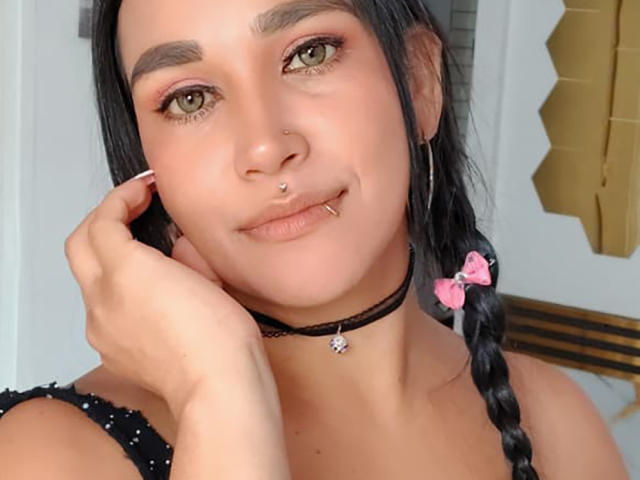 EdenIvyLm, con trenzas negras, un clip rosa con forma de mariposa, un aro en la nariz, un piercing en el labio, aros grandes, un collar negro y maquillaje, sonriendo y con la mano en la mejilla. Tiene ojos marrones claros..