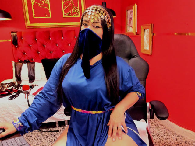 Anissa Ali, con i suoi lunghi capelli neri, indossa un abito di seta blu, un copricapo dorato e una maschera nera, seduta su una sedia con le gambe divaricate, in una stanza rossa. Il suo sedere è visibile..