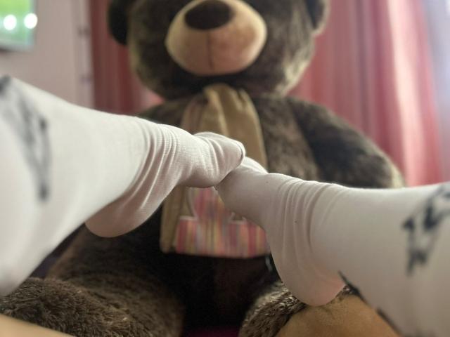 As meias brancas de Katie Kingston estão em contato com o pé de um enorme urso de pelúcia marrom em um momento suave, fofo, íntimo e ligeiramente divertido. O nariz do urso está ligeiramente borrado ao fundo..