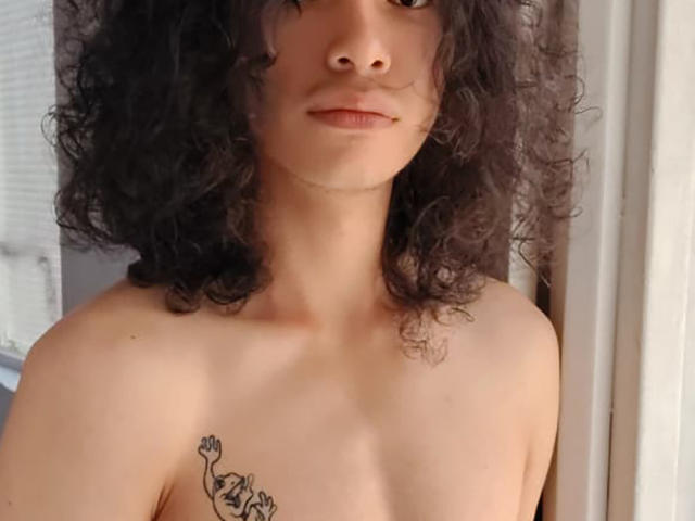 FrankieManHot, con capelli scuri e ricci, pelle chiara e un piccolo tatuaggio sul lato sinistro del petto, in piedi senza maglietta, mostrando le spalle nude e un'espressione neutra..
