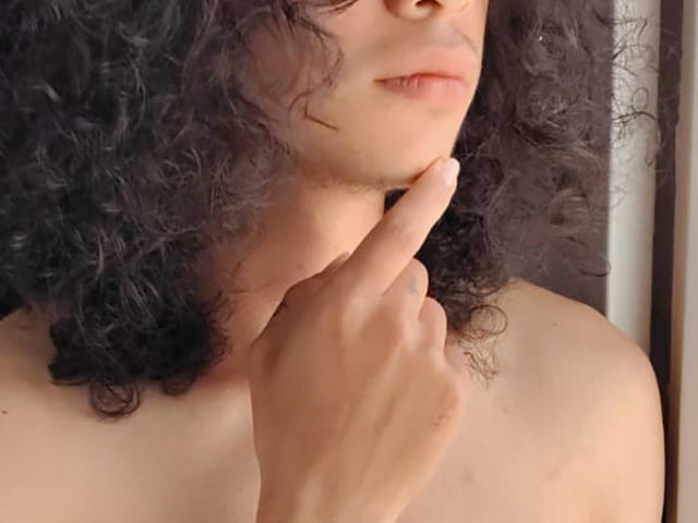 FrankieManHot, avec ses cheveux noirs bouclés, sa peau claire et ses lèvres roses pâles, touche son menton avec un doigt, laissant apparaître ses épaules nues. Sa main est près de son cou, la touchant légèrement..