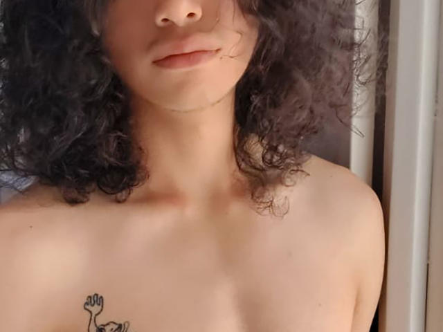 FrankieManHot, com cabelo escuro e encaracolado, pele clara e uma pequena tatuagem de uma mão no peito. Seu corpo superior nu revela uma aparência suave e natural..