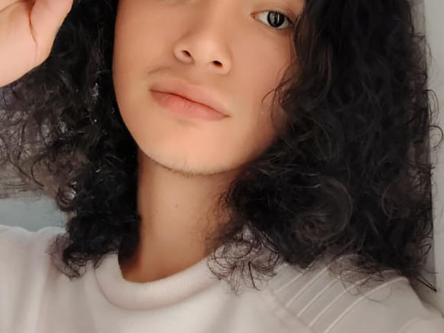 FrankieManHot tiene el pelo negro y rizado, piel de color marrón claro, y lleva una camisa blanca, con labios rosados ligeramente separados y un pequeño bigote. Los ojos de FrankieManHot están mirando hacia arriba..