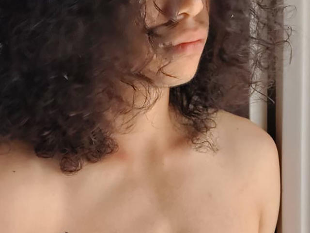 FrankieManHot, con cabello oscuro y rizado, piel pálida y una sonrisa suave, mostrando hombros desnudos y un pecho sutil. Sin ropa, solo piel lisa y belleza natural..