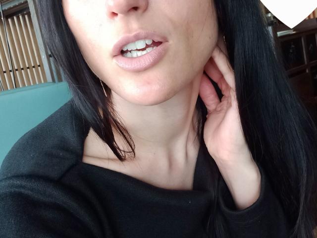 Il viso di BellaJulls, con i capelli neri, le labbra rosa leggermente schiuse che mostrano i denti, e una mano che tocca il collo. Indossa una maglia nera che mette in risalto la clavicola e la parte superiore del petto..