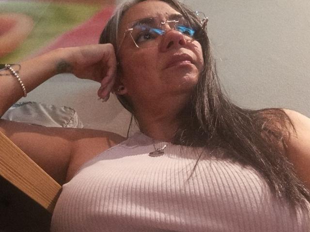 LailaCastillo with glasses, pink ribbed top, large tits, arm raised, bracelet on wrist, looking up, brown hair, light skin, subtle makeup, relaxed, slight smile, indoor light.