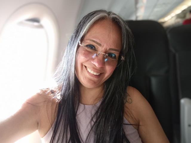 LailaCastillo has long black and white hair, glasses, and a cute smile, sitting in a plane with light coming from a window on her right. Her tits are small.