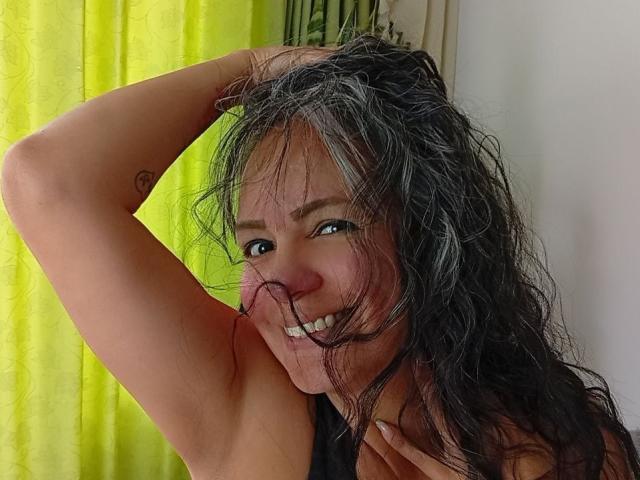 LailaCastillo with wavy black hair, holding her arm up, smiling, showing her tits, green curtain in the background, small heart tattoo on her arm.